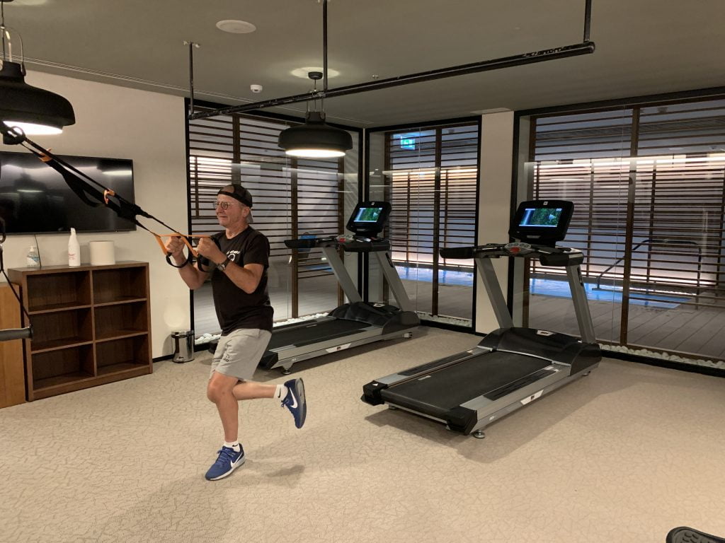 L'image montre un homme dans une salle de sport intérieure bien éclairée. L'homme porte des vêtements décontractés et des baskets, et il utilise une sorte de sangle de suspension pour faire de l'exercice. Derrière lui, il y a deux tapis roulants avec des écrans intégrés. La salle est moderne avec un éclairage suspendu, des stores horizontaux aux fenêtres, et un sol de salle de sport claire. Il y a également quelques meubles et accessoires, comme une étagère et une bouteille. La pièce a l'air spacieuse et bien aménagée pour divers types d'entraînements.